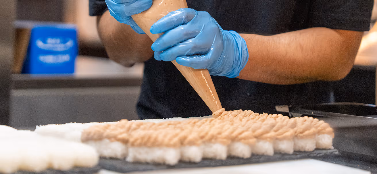 Gloved hands piping topping onto rows of sushi.